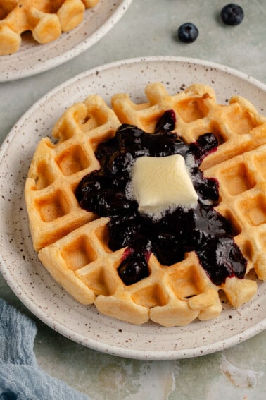 golden waffle on white plate with blueberry compote and melting pat of butter