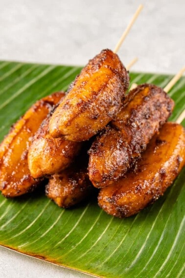 a photo of caramelized bananas on barbecue sticks laying on a banana leaf