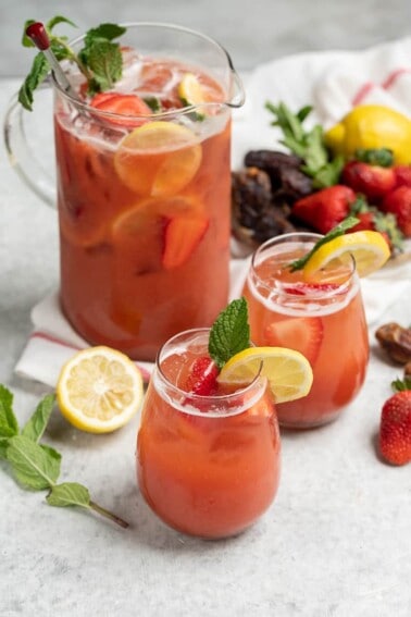 two glasses of strawberry lemonade on white table with full pitcher in the background