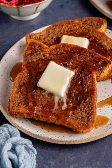 white plate with 2 slices of vegan gingerbread french toast with a square of melting butter, maple syrup and powdered sugar