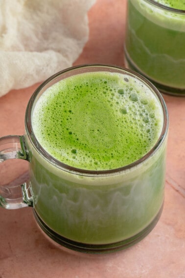 2 frothy matcha lattes in glass cups on pink table with white towel